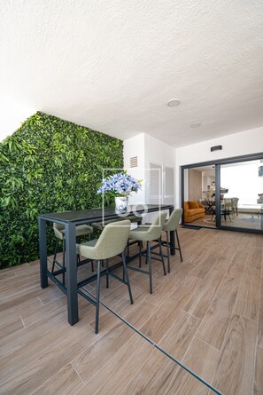 A modern dining area with a dark wooden table and chairs, a green leafy wall, and a view into a kitchen area. - Casa de Adamant - Ground floor resort apartment with BBQ, shared jacuzzi and pool by OYOOSTAYS (Torrevieja)