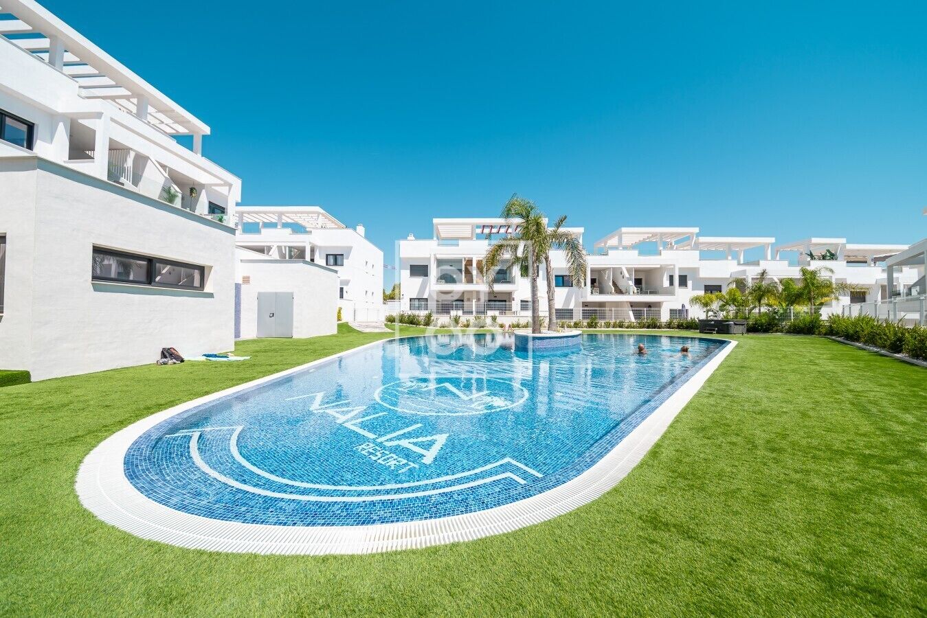 A modern pool area with a clear blue pool, surrounded by well-maintained grass and white buildings.
