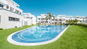 Un área de piscina moderna con una piscina azul cristalina, rodeada de césped cuidado y edificios blancos.