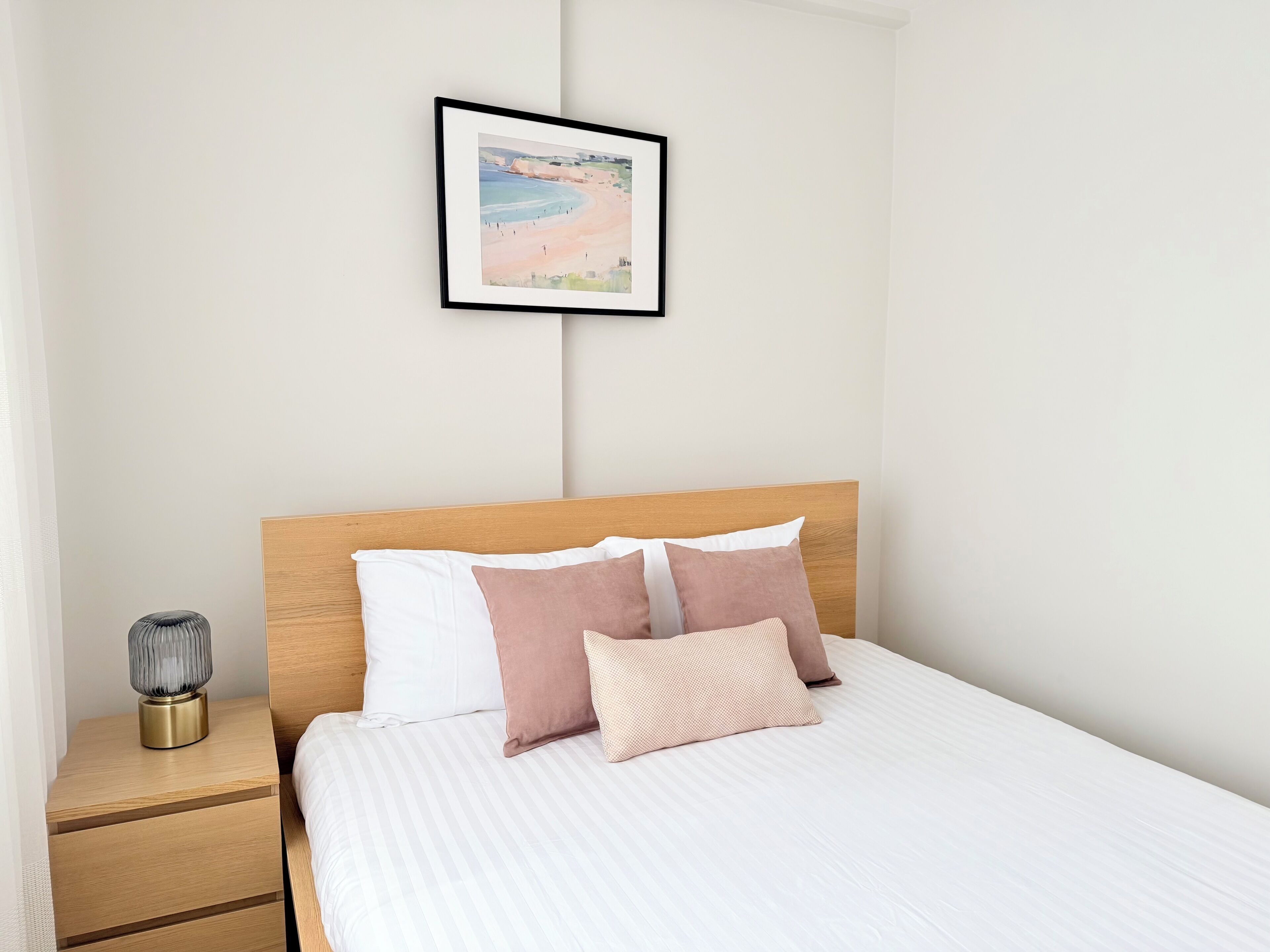Un lit bien fait, avec une tête de lit en bois, une table de chevet avec une lampe et, au mur, un tableau représentant une scène de plage.