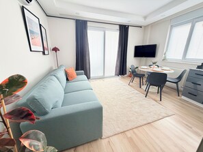 A modern living room with a sofa, a dining table with chairs, a TV mounted on the wall, and large windows with curtains. - Modern Central Flat Near Old Town with Fast WiFi 2BR (ANTALYA)