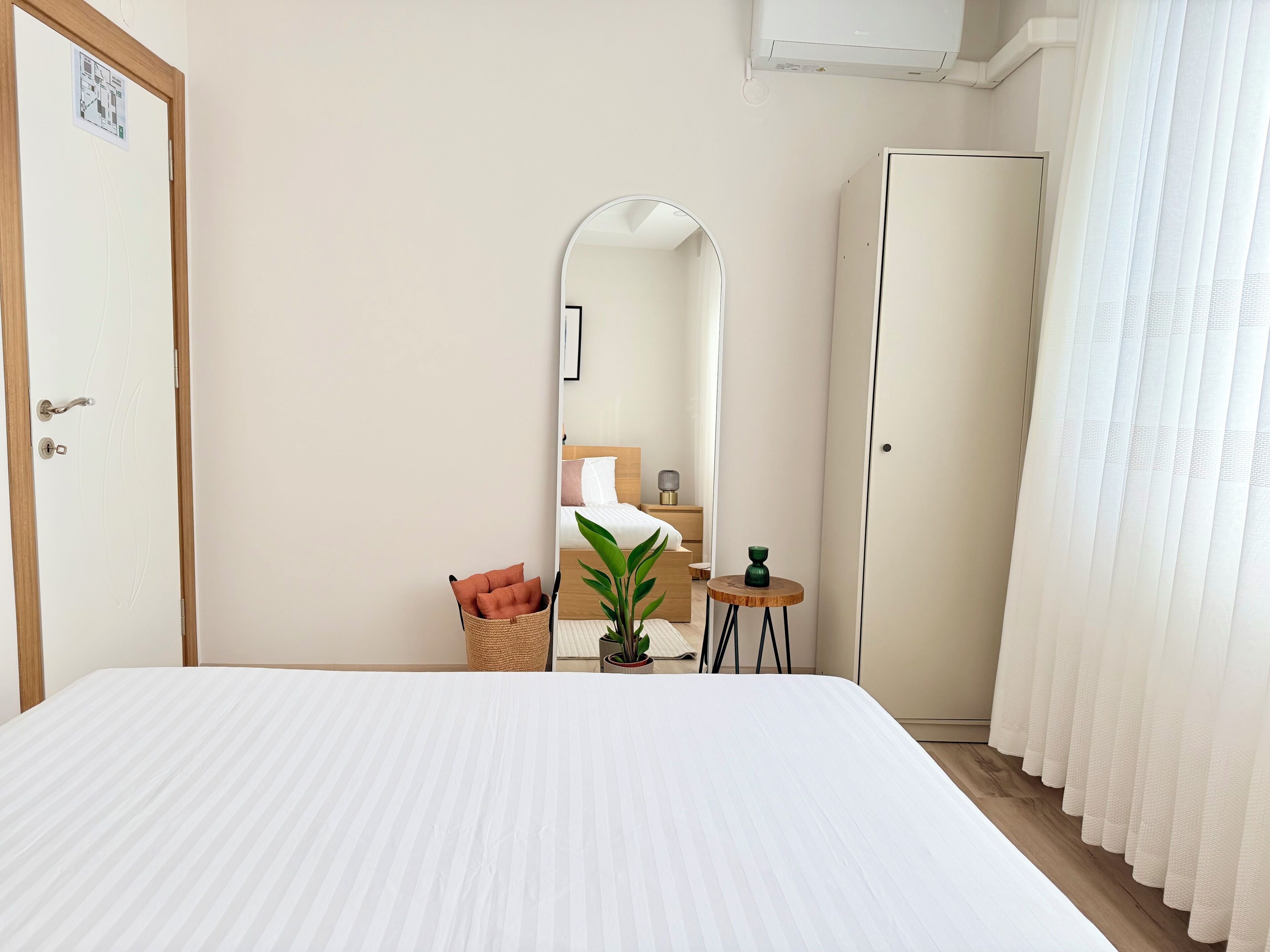 A hotel room with a large bed, a wardrobe, a mirror, a small table with a plant, and a window with curtains.