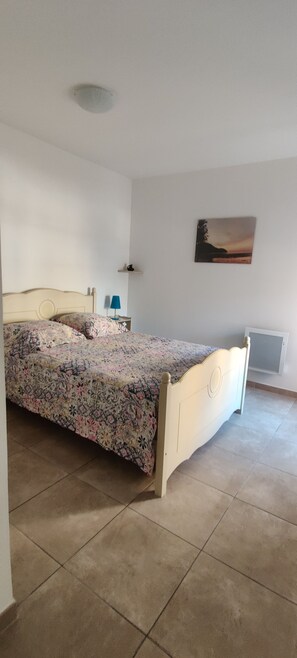 A bedroom with a bed, a nightstand, a lamp, a wall-mounted picture, and a radiator. - Beautiful 3-Bedroom Apartment in the Village Center in a New Building (Solliès-Pont)