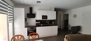 A compact kitchen with white cabinets, a black countertop, and a microwave. There is a dining area with a table and chairs. - Beautiful 3-Bedroom Apartment in the Village Center in a New Building (Solliès-Pont)