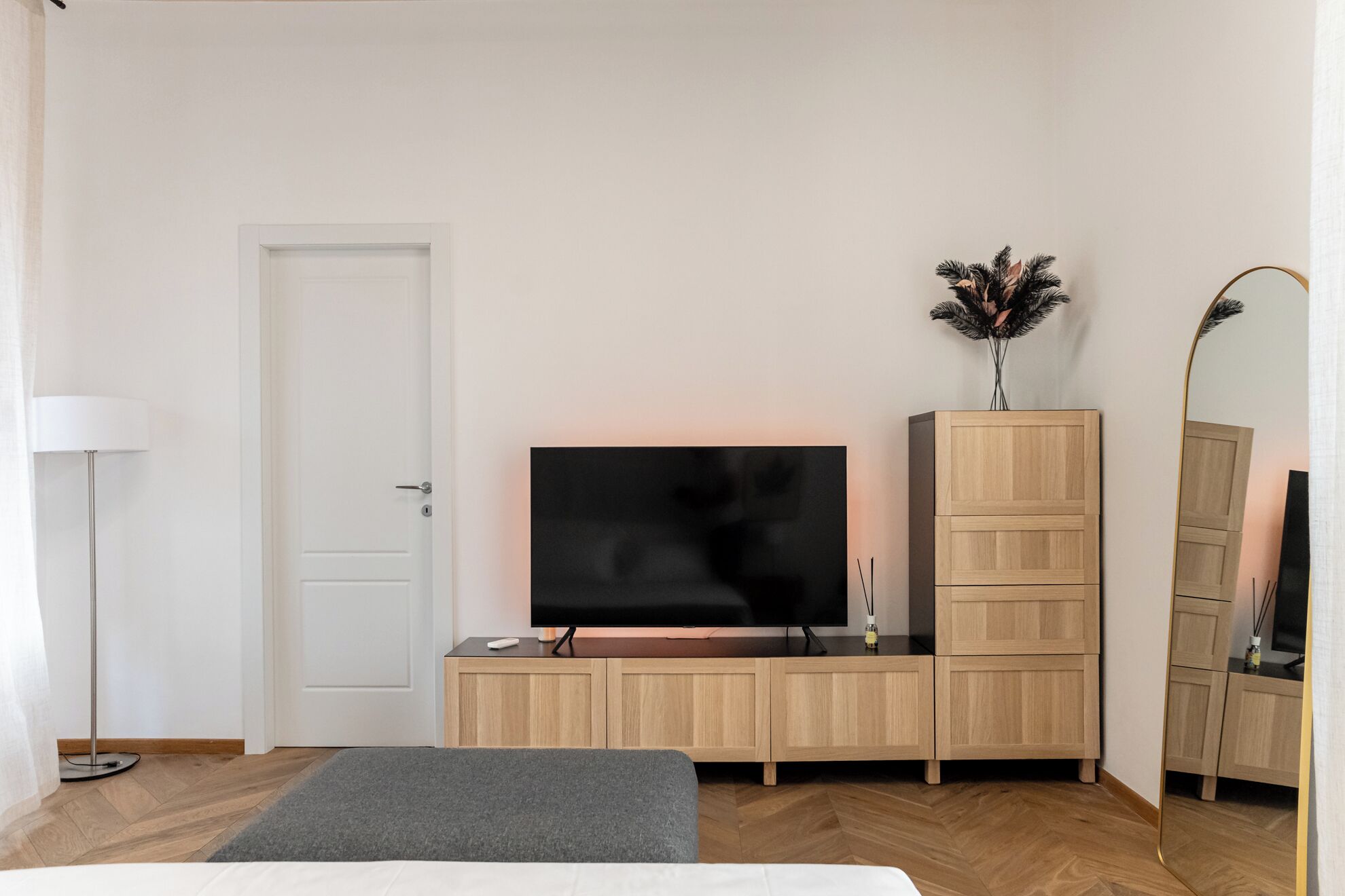 Una camera da letto con un letto, una TV su un mobile in legno, uno specchio e una lampada da terra.