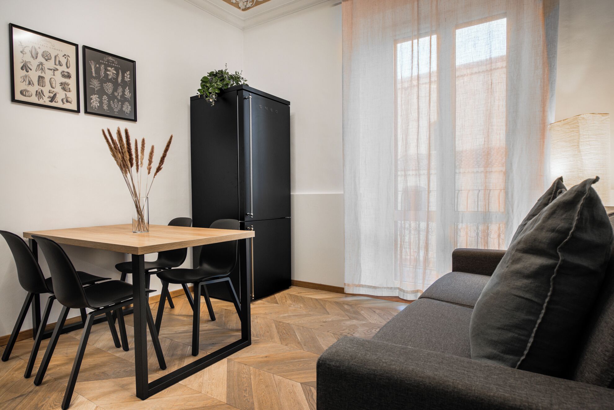 A modern living space with a dining table, chairs, a sofa, a black refrigerator, and a window with sheer curtains.