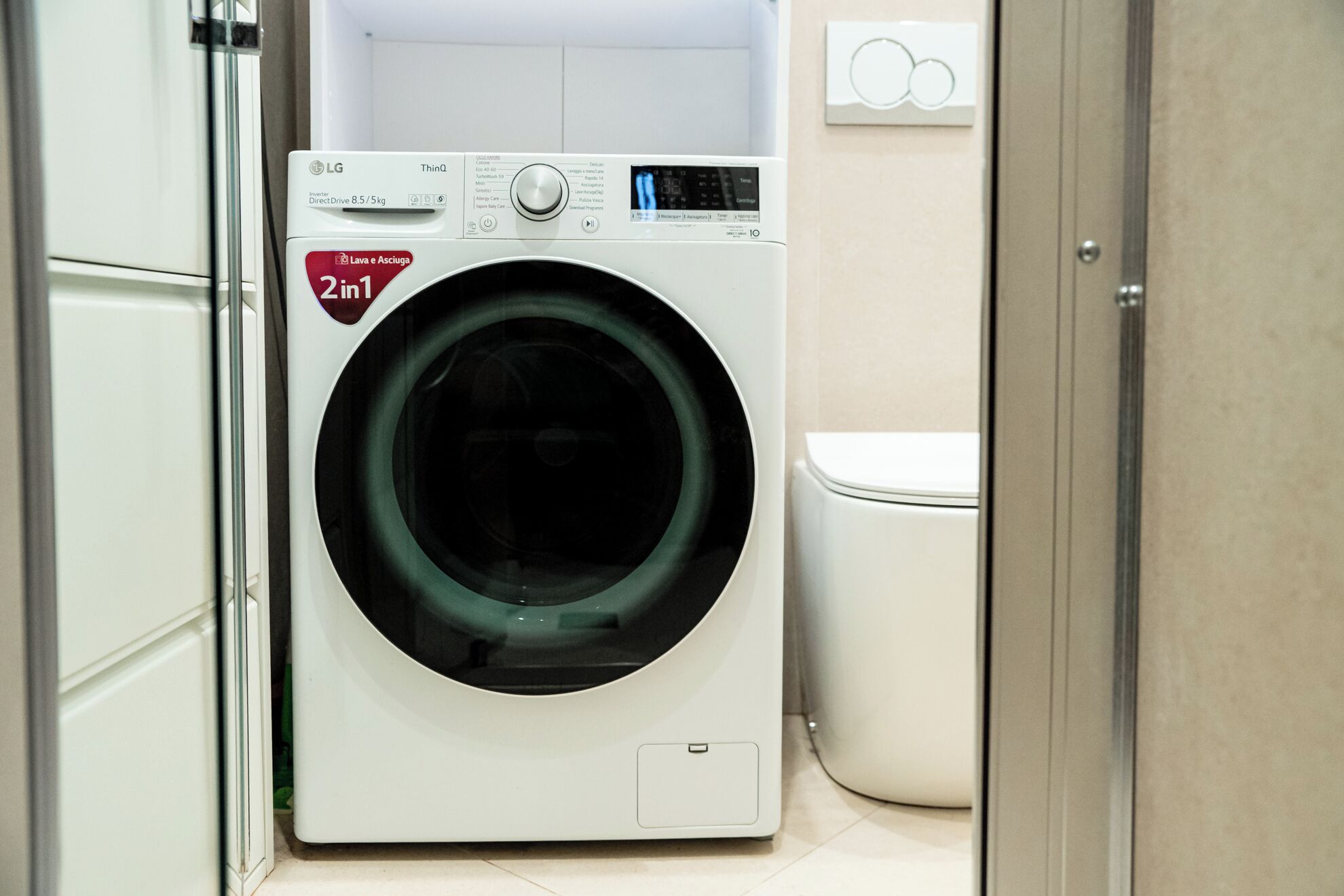Una lavatrice a carica frontale con pannello di controllo, un water bianco e una porta che conduce in un'altra stanza.