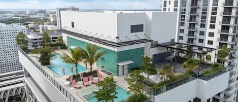 Uma piscina no terraço com espreguiçadeiras e palmeiras, cercada por edifícios altos.