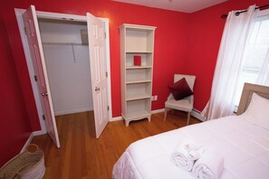 Ein Schlafzimmer mit roter Wand, einem weißen Bett, einem weißen Kleiderschrank und einem Sessel mit rotem Kopfkissen.