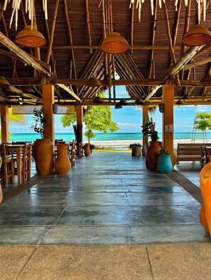 An open-air dining area with a view of the beach, featuring a thatched roof, wooden furniture, and potted plants.
