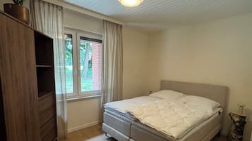 Ein Schlafzimmer mit einem Bett, einer Holzkommode, einem Fenster und einer Deckenleuchte.