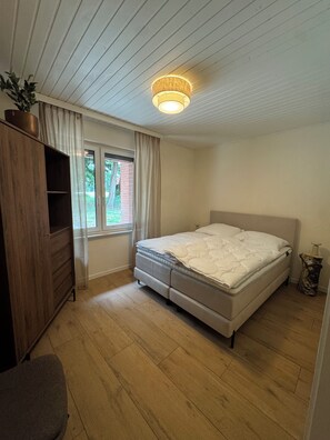 Ein Schlafzimmer mit einem Bett, einer Holzkommode, einem Fenster und einer Deckenleuchte.