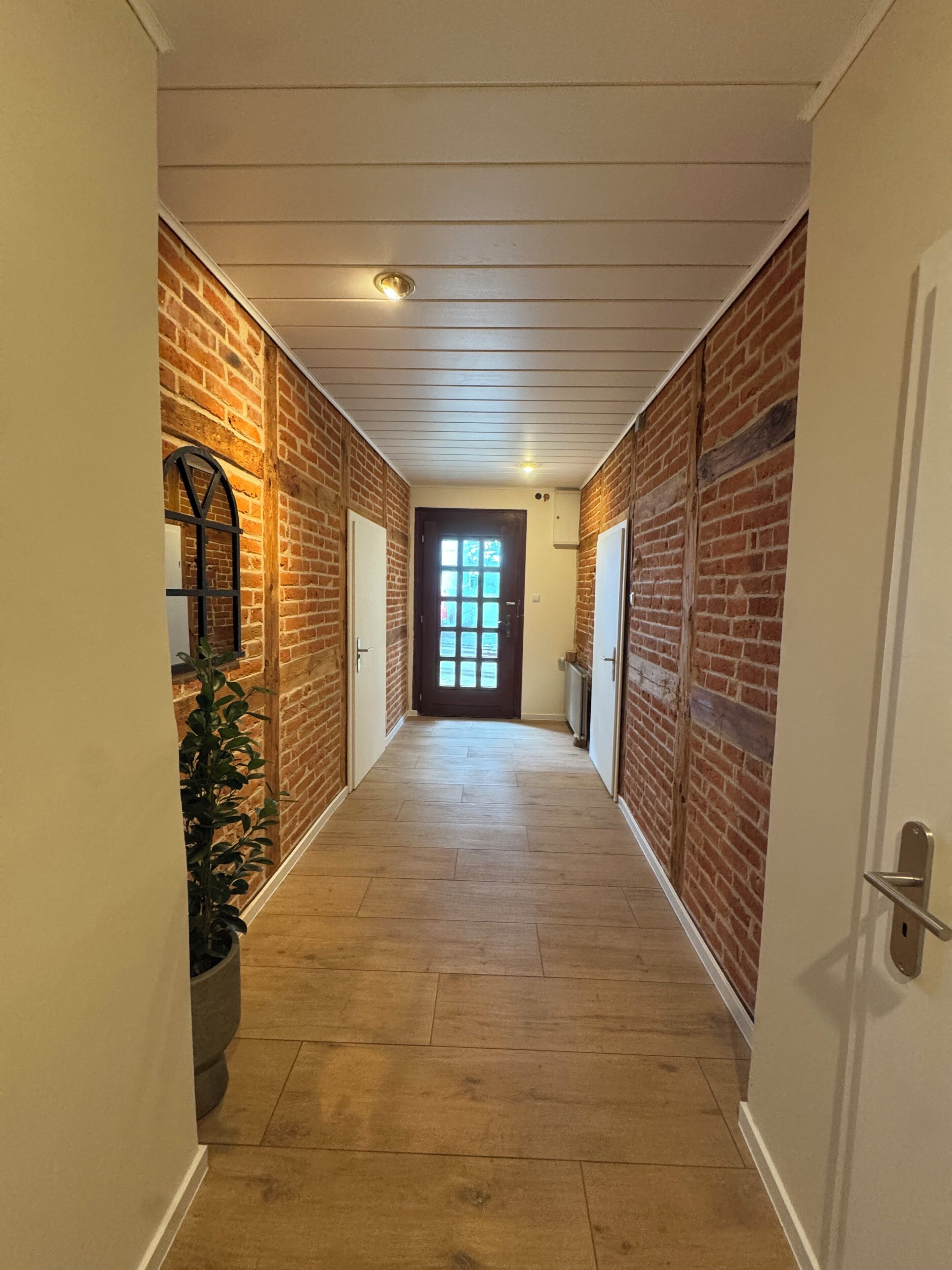 Un couloir aux murs en briques, avec un plancher en bois et une porte au fond.