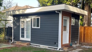 A single-story house with a dark siding exterior, white trim around windows and doors, a small porch, and a wooden fence.