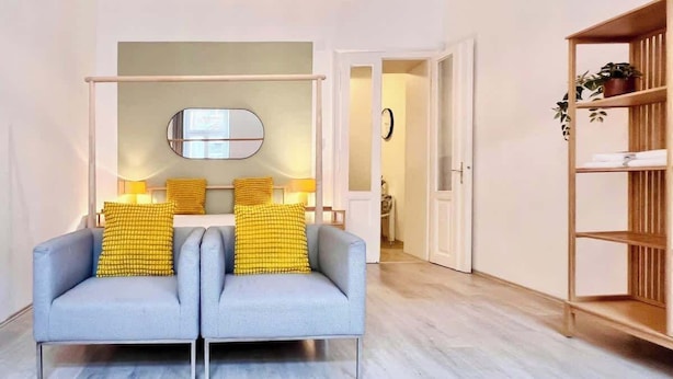 Ein modernes Wohnzimmer mit einem grauen Sofa, zwei gelben Kissen, einem Holzregal und einem Spiegel an der Wand.