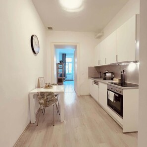 A modern kitchen with white cabinets, a stainless steel stove, and a dining area with a white table and patterned chair. - Servus Homes - Metro Stadthalle - Amadeus (Vienna)