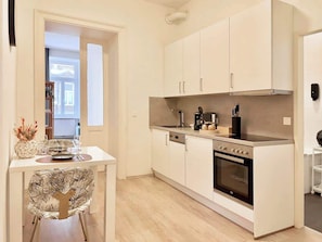 A compact kitchen with white cabinets, a built-in oven, and a dining area with a table and chairs. - Servus Homes - Metro Stadthalle - Amadeus (Vienna)