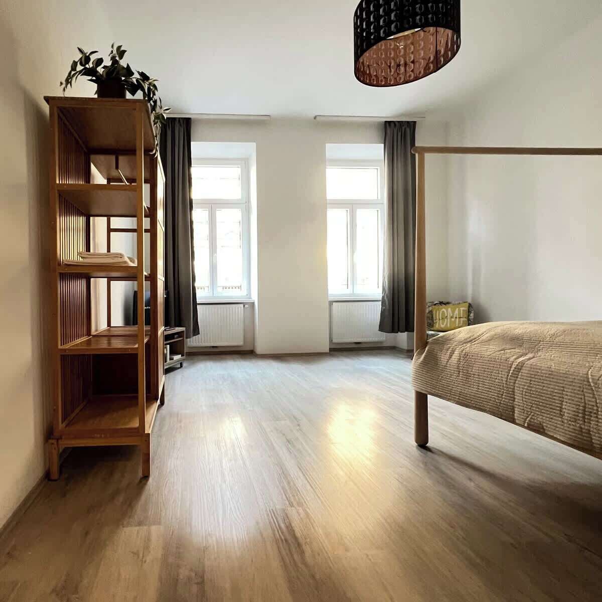 A bedroom with a wooden floor, a bed, a bookshelf, and a window with curtains.