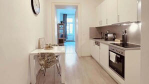 A modern kitchen with white cabinets, a stainless steel stove, and a dining area with a white table and patterned chair. - Servus Homes - Metro Stadthalle - SISSI (Wien)