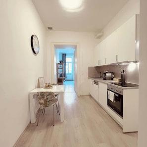A modern kitchen with white cabinets, a stainless steel stove, and a dining area with a white table and patterned chair. - Servus Homes - Metro Stadthalle - SISSI (Wien)