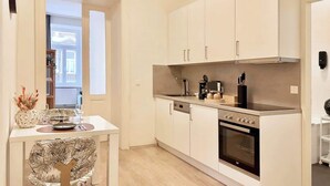 A compact kitchen with white cabinets, a built-in oven, and a dining area with a table and chairs. - Servus Homes - Metro Stadthalle - SISSI (Wien)