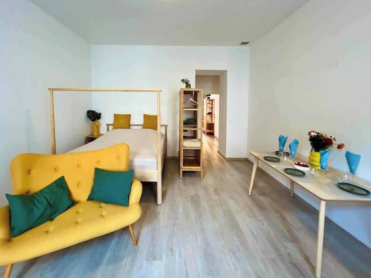 Een kamer met een gele bank, een bed met houten frame, een houten ladder en een tafel met borden en glazen.