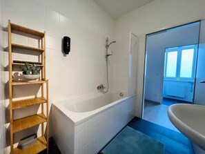 A bathroom with a bathtub, a wooden shelf with plants, a hand dryer, and a view of the outside. - Servus Homes - Metro Stadthalle - Vienna Calling (Wien)