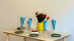 Une table à manger avec des serviettes bleues, un vase jaune rempli de fleurs et un saladier de fruits.