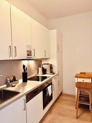 Een moderne keuken met witte kastjes, een ingebouwde oven, een koffiezetapparaat en een houten eettafel met een stoel.