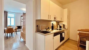 A modern kitchen with white cabinets, a built-in oven, and a dining area with a table and chairs. - SOUND OF MUSIC · Servus Homes – Metro Stadthalle – Sound Of Music (Vienna)