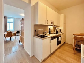 A modern kitchen with white cabinets, a built-in oven, and a dining area with a table and chairs. - SOUND OF MUSIC · Servus Homes – Metro Stadthalle – Sound Of Music (Vienna)