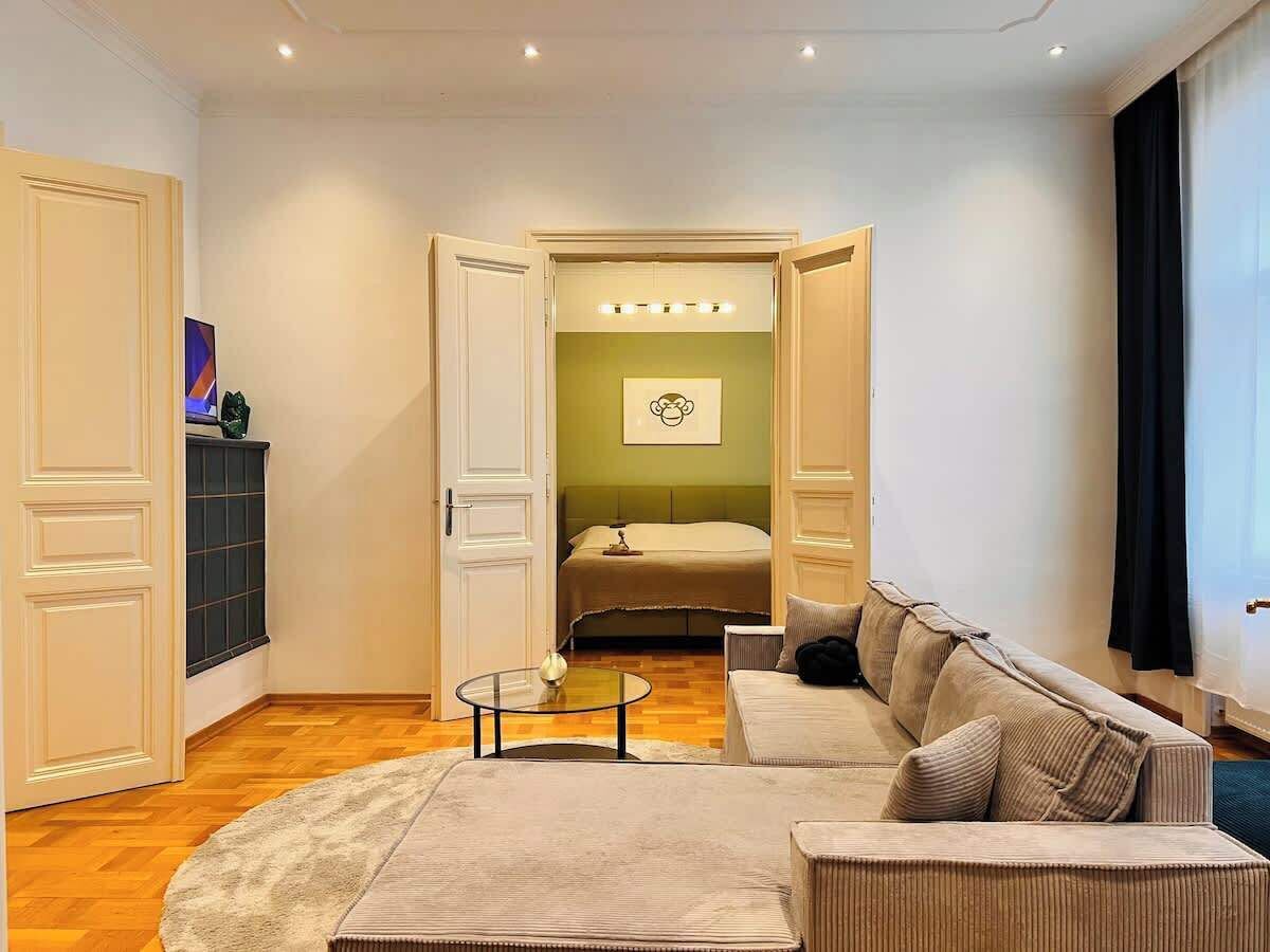 Ein Hotelzimmer mit einer Couch, einem runden Couchtisch, einem Bett und einer Wand in Grünton sowie einem Fenster mit Vorhängen.