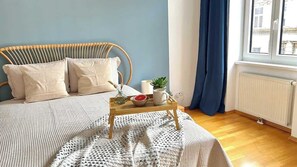 A bed with a wooden headboard, a bedside table with a bowl and a plant, and a window with a view of buildings. - Servus Homes - Metro Stadthalle - FRANZ (Wien)