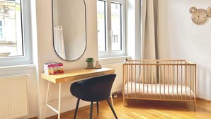 Ein Zimmer mit Holzboden, einem Schreibtisch mit Stuhl, einem Babybett, einem Fenster mit Vorhängen und einem Heizkörper.