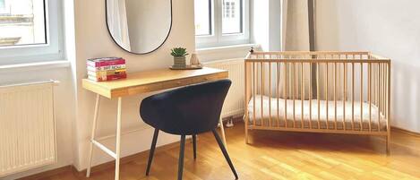 A room with a wooden floor, a desk with a chair, a crib, a window with curtains, and a radiator.