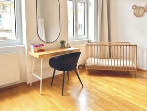 Ein Zimmer mit Holzboden, einem Schreibtisch mit Stuhl, einem Babybett, einem Fenster mit Vorhängen und einem Heizkörper.
