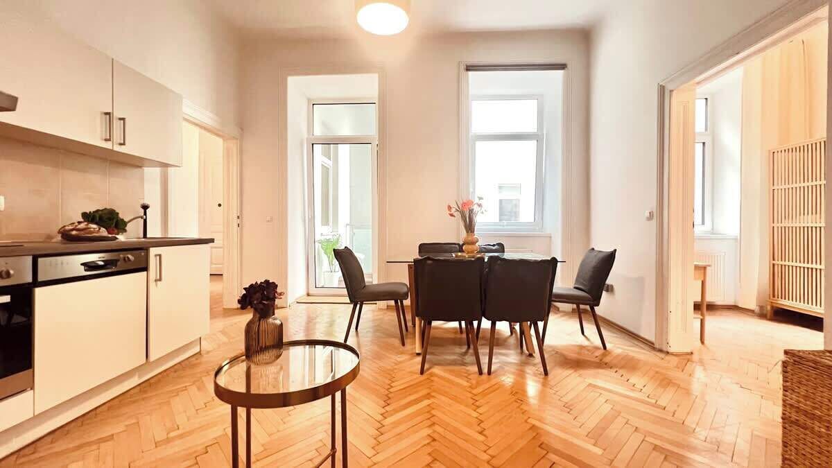 Uma cozinha moderna com armários brancos, uma área de jantar com mesa redonda e quatro cadeiras, e uma janela grande que oferece luz natural.