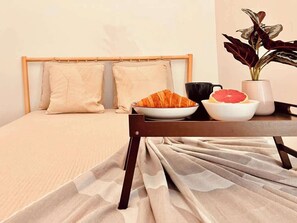 Ein ordentlich bezogenes Bett mit Kopfteil, ein Tisch mit einer Schüssel Essen, einer Tasse und einer Pflanze.