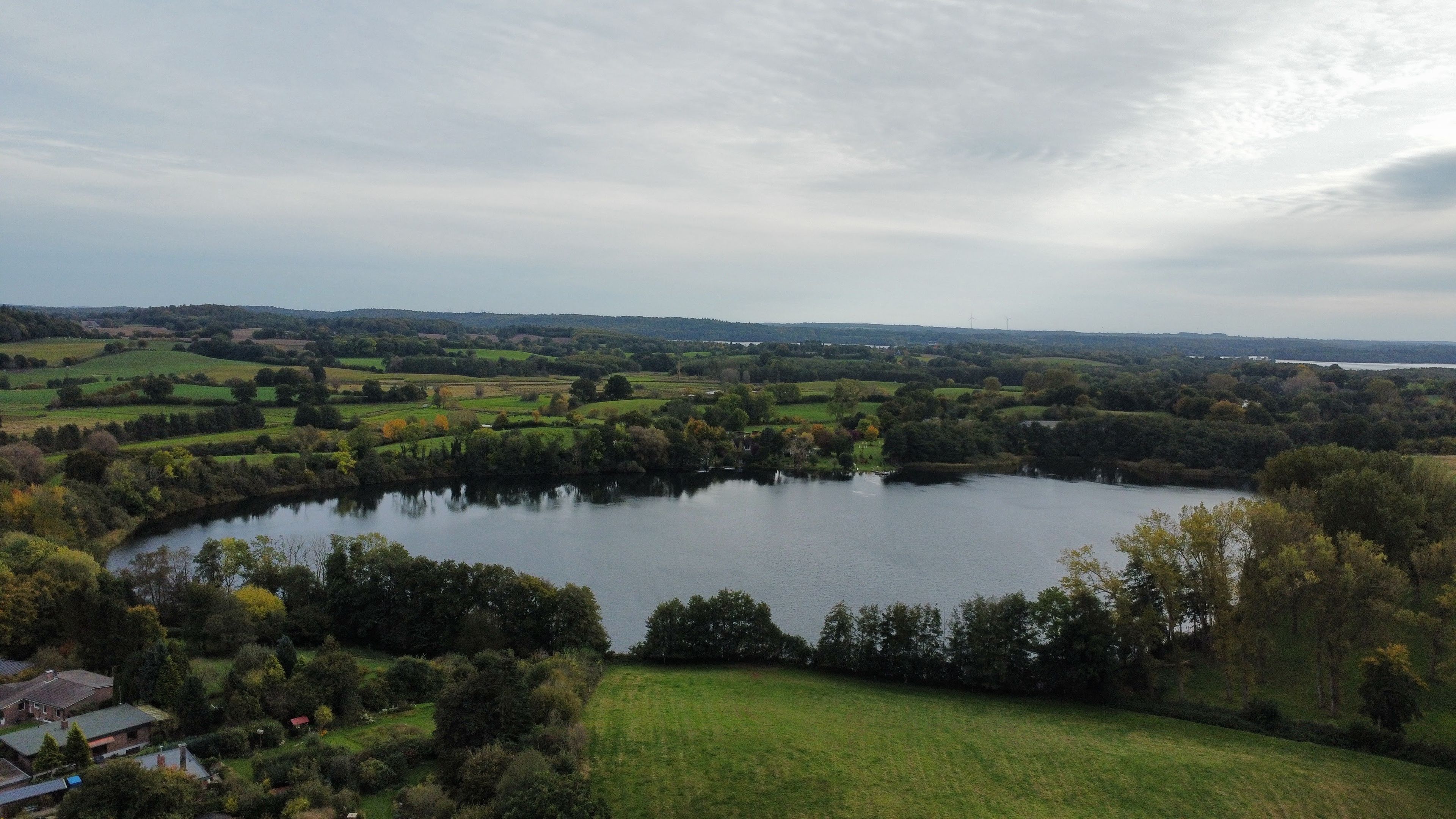 Un llac envoltat per camps verds i arbres, amb alguns edificis al primer pla.