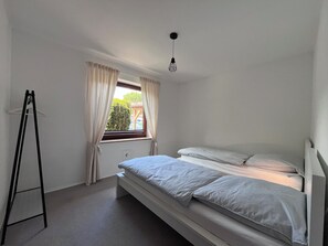 A small, single-bed room with a window, a hanging light fixture, and a black metal stand.