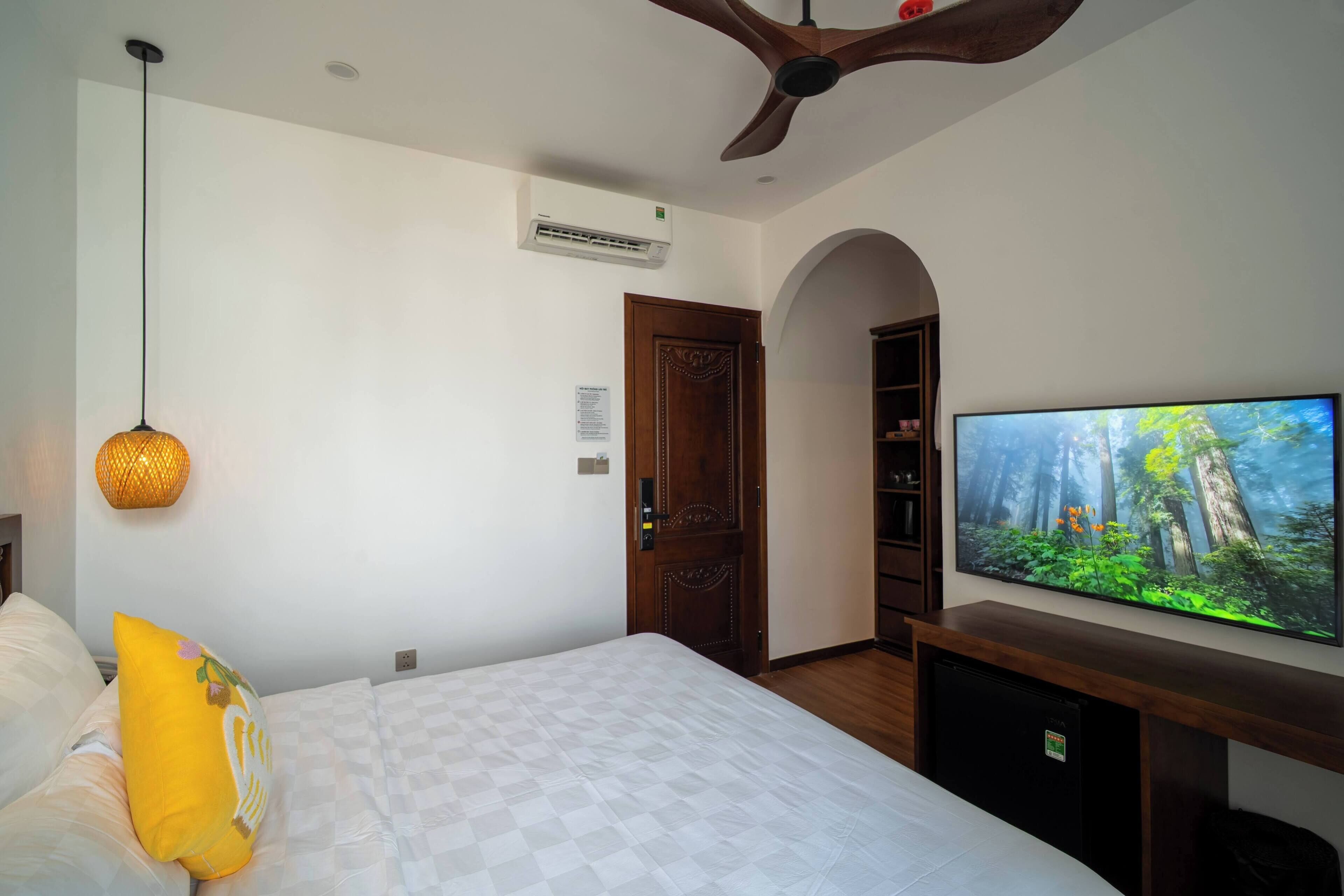 Quarto com cama, televisão, ventoinha no teto, aparelho de ar condicionado e porta de madeira.