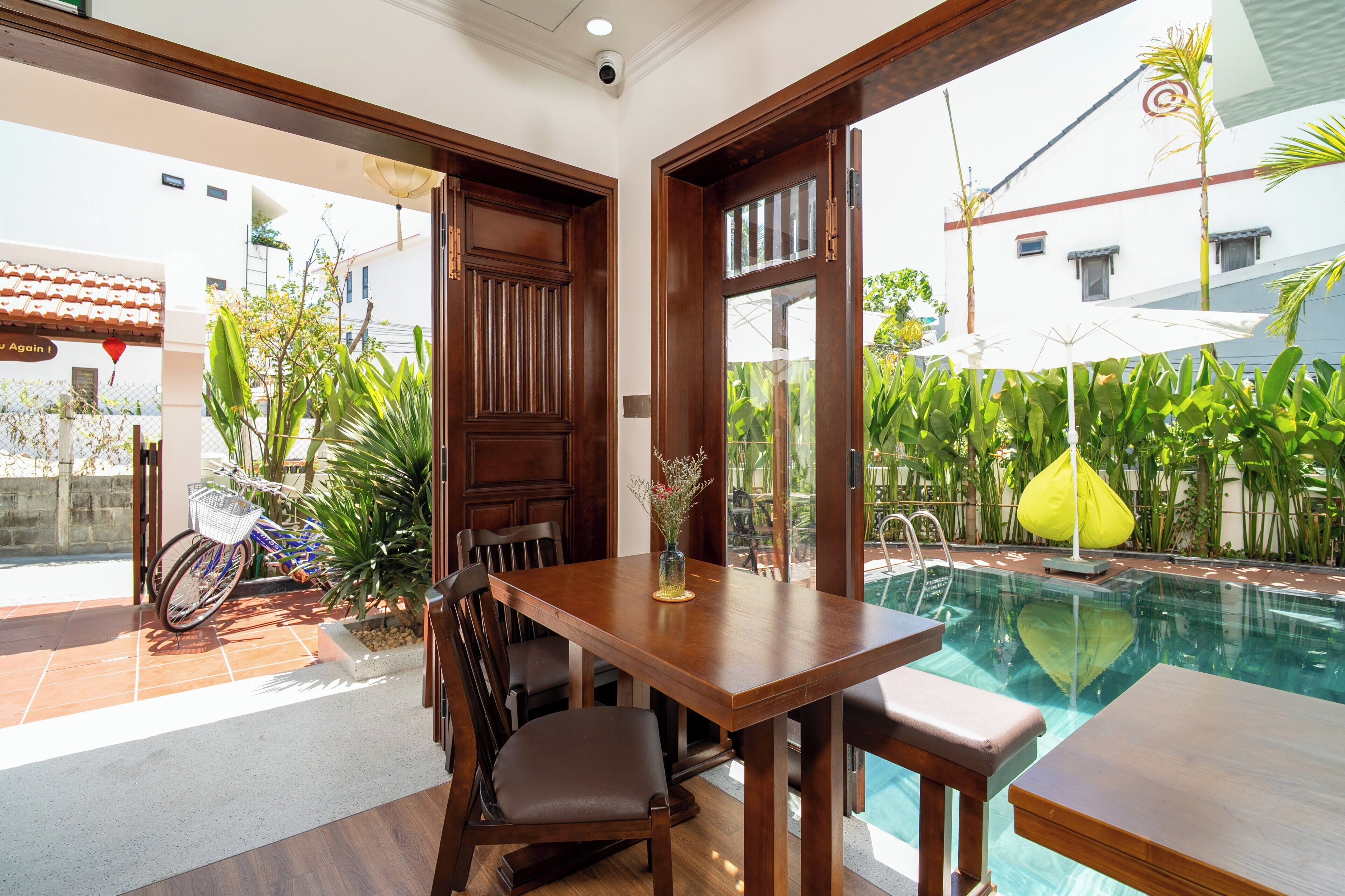 A poolside area with a wooden table and chairs, a glass-enclosed door, and a view of the pool and surrounding greenery.