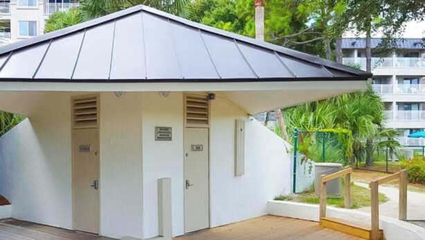 Une petite construction blanche, composée d’une seule pièce, avec un toit en pente, une porte et une fenêtre, entourée de verdure et dotée d’une terrasse en bois.