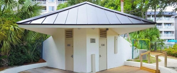 Ein kleines, weißes Gebäude mit einem Zimmer, schrägem Dach, einer Tür und einem Fenster, umgeben von Grün und einer Holzterrasse.
