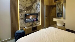 A modern hotel room with a large bed, a TV mounted on a marble wall, a desk with a chair, and a wardrobe.
