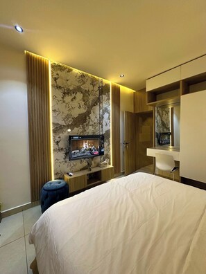 Una habitación de hotel moderna con una cama grande, un televisor montado en una pared de mármol, un escritorio con una silla y un armario.