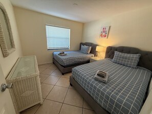 A bedroom with two beds, a window with blinds, a bedside lamp, and a decorative wall piece. - Sunny 2BR Condo w/ Heated Pool — Minutes to IMG Academy & Beaches (Bradenton)