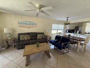 Ein Wohnzimmer mit Sofa, Couchtisch und Sesseln, einem Esstisch mit Stühlen, einem Deckenventilator und einer Küchenzeile.
