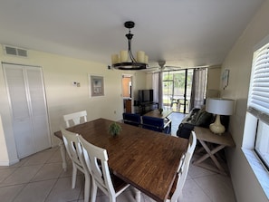 A dining area with a wooden table and white chairs, a sofa, a TV, and a window with blinds. - Sunny 2BR Condo w/ Heated Pool — Minutes to IMG Academy & Beaches (Bradenton)