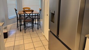 Una cocina con un refrigerador de acero inoxidable, una mesa de comedor de madera con cuatro sillas y una ventana con persianas.